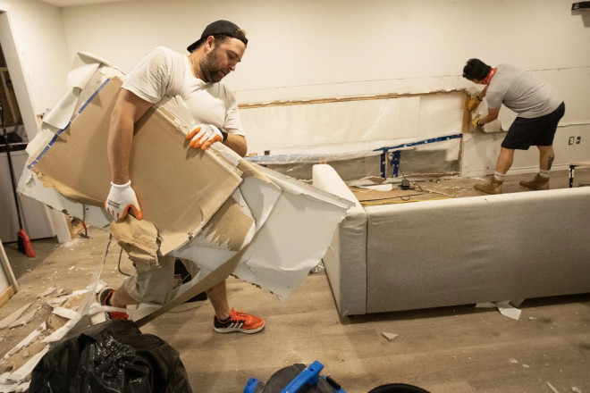 A man carries a stack of damaged drywall as another man pulls pieces from the wall in the background.